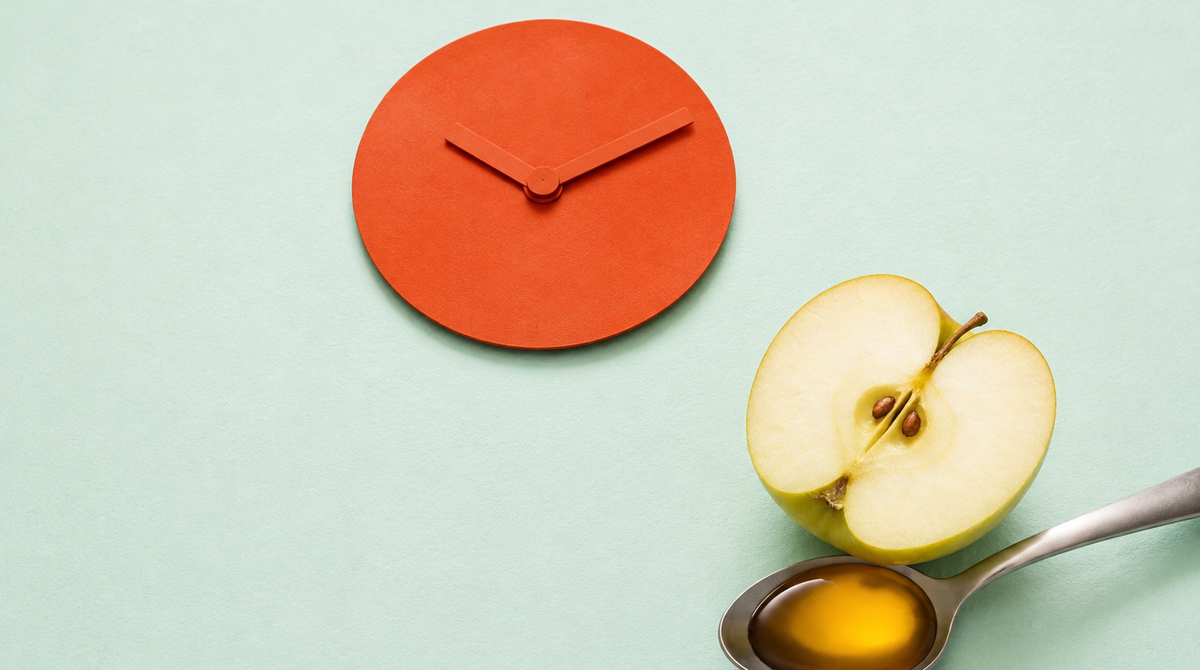 A halved apple and a stainless spoon of amber apple cider vinegar beside a coral paper clock face on mint paper